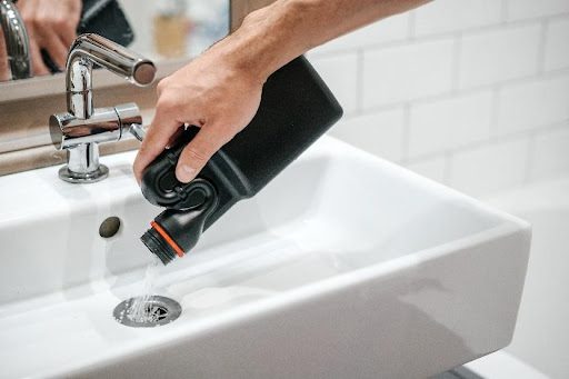 A homeowner pouring a black plastic container filled with harmful chemicals into the drain to clear a clog that needs professional plumbers in St. Louis, MO.