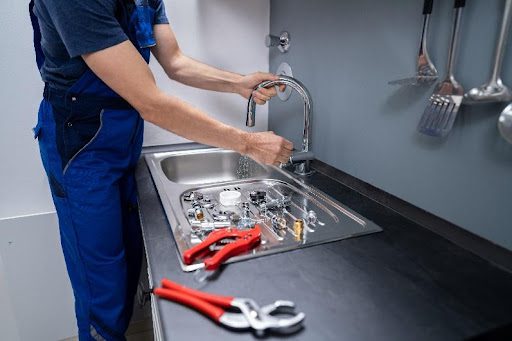 A plumber in Bridgeton, MO, wearing blue overalls and fixing a plumbing emergency in a residential kitchen with professional tools.