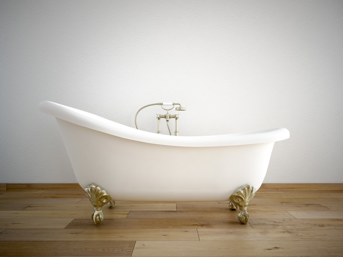 A white clawfoot bathtub with brass fixtures is centered against a plain white wall on a wood floor.