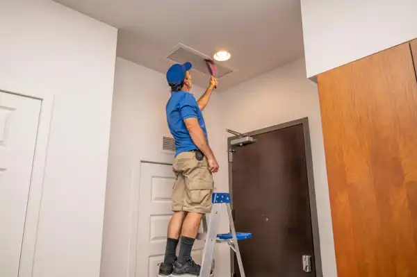 A person in a blue shirt and cap stands on a ladder, meticulously performing air duct cleaning by brushing the ceiling vent.