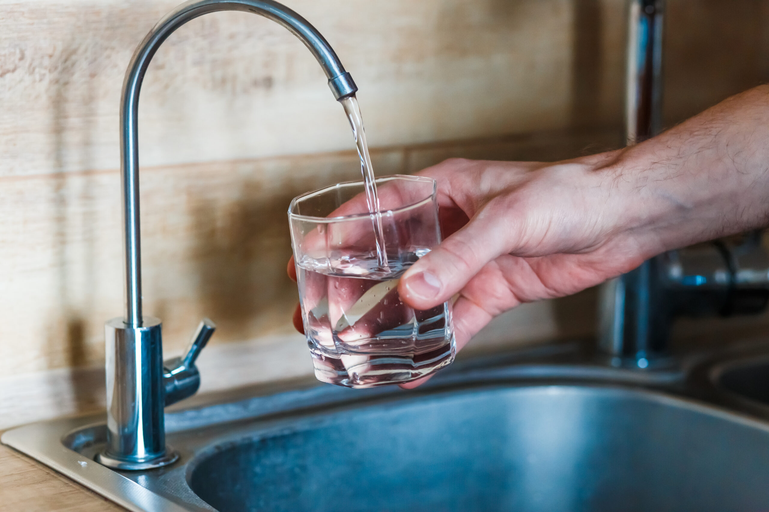 A person fills a glass with water from a kitchen faucet above a stainless steel sink.